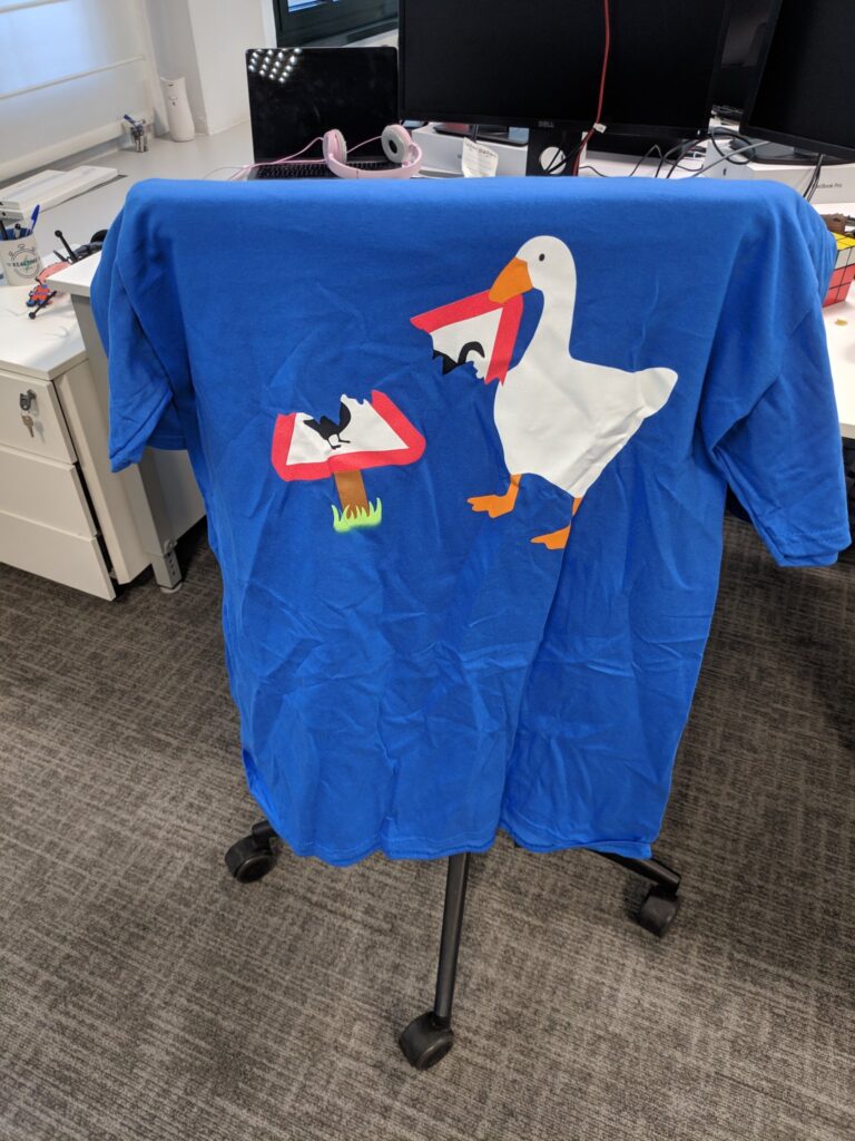Foto de unha camiseta apoiada sobre o respaldo de unha cadeira de oficina. A camiseta é azul e ten unha imaxe dun ganso rompendo un sinal de tráfico de "perigo: ganso". É unha imaxe promocional do xogo Untitled Goose Game.